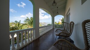 A covered balcony with white railings, wicker chairs, and a view of palm trees and buildings. - The Ambassador Full Building (Jan Thiel)