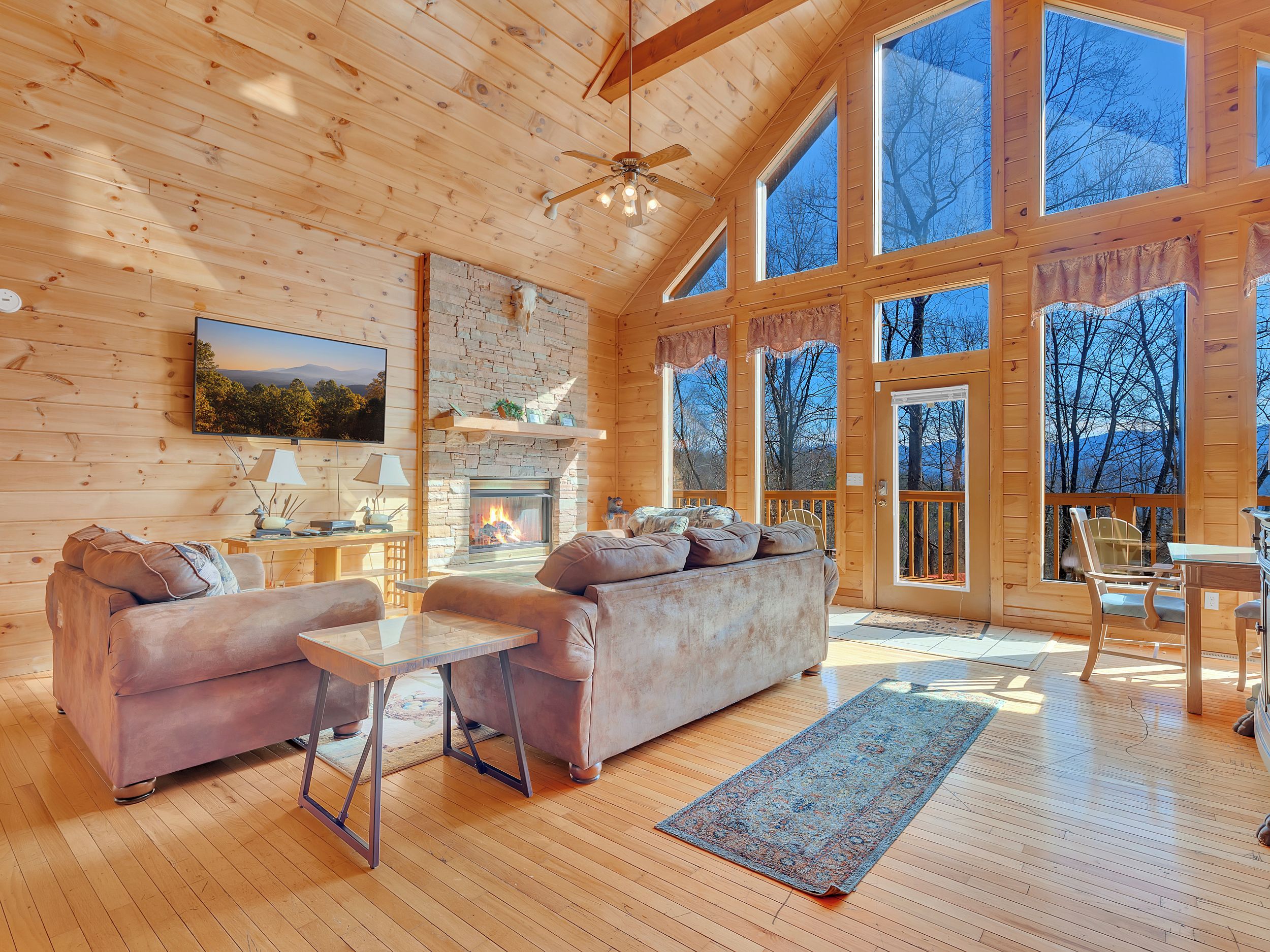 A cozy cabin interior with a fireplace, wooden walls, large windows, and a comfortable seating area.