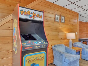 Une borne d’arcade Pac-Man dans une pièce aux murs en bois, avec un fauteuil bleu et une lampe.