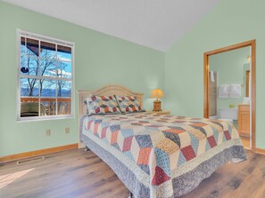 Une chambre à coucher comprenant un lit, une table de chevet, une fenêtre donnant sur des arbres et des montagnes, et une porte menant à une salle de bain.