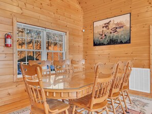 Interior de una cabaña de madera con una mesa redonda de vidrio rodeada de sillas de madera, un ventanal con vistas a árboles y un cuadro de pájaros en la pared.