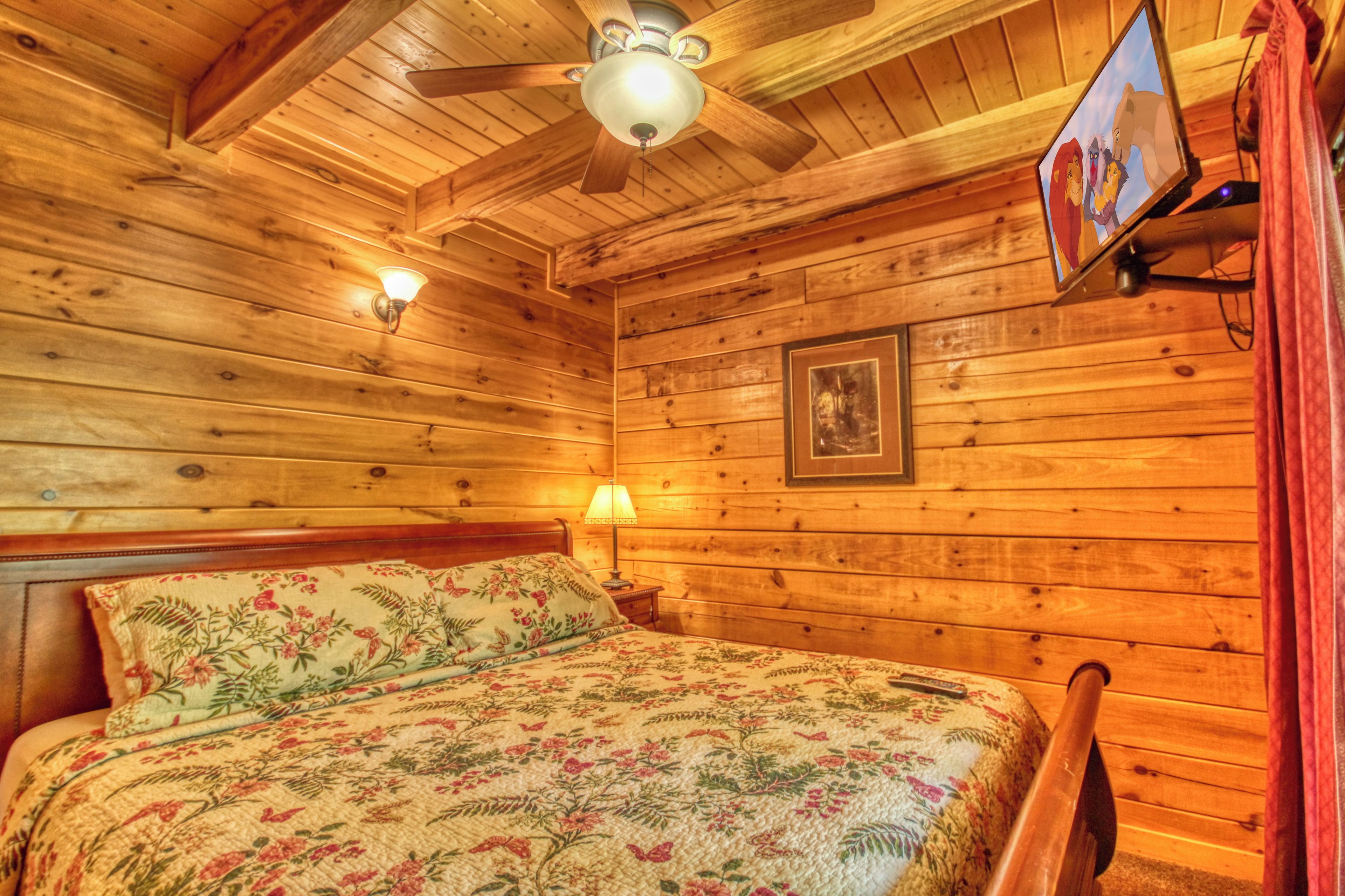 Ein Zimmer einer Holzhütte mit einem Bett, einem Deckenventilator, einem an der Wand befestigten Fernseher und einem gerahmten Bild.