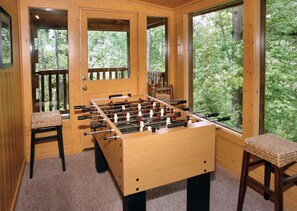 Ein Zimmer mit Holzboden, einem Kicker, zwei Stühlen und großen Fenstern mit Blick auf Bäume.