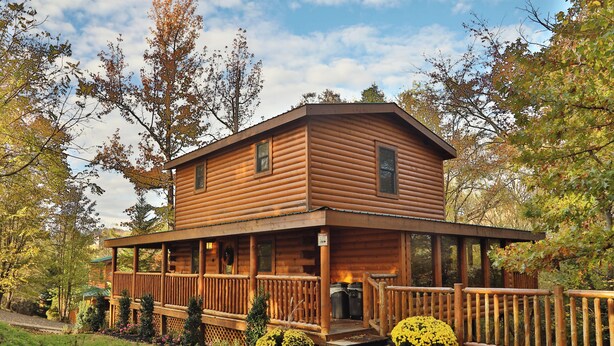 Eine Holzblockhütte mit Veranda und umzäunter Terrasse, umgeben von Bäumen und Sträuchern.