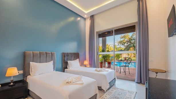 A hotel room with two beds, a TV, a desk, and a view of an outdoor pool area. - Dar Fatima Tamazouzt (Tamazouzte)