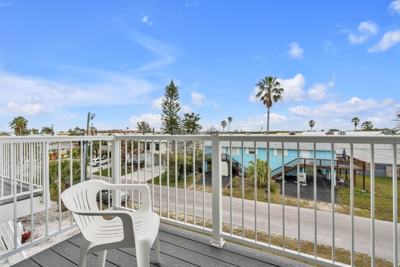 Un balcon avec une chaise en plastique blanche, une rambarde en métal blanc et une vue sur un immeuble doté d’une piscine et de palmiers.