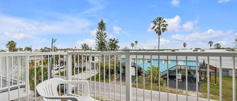 Un balcon avec une chaise en plastique blanche, une rambarde en métal blanc et une vue sur un immeuble doté d’une piscine et de palmiers.