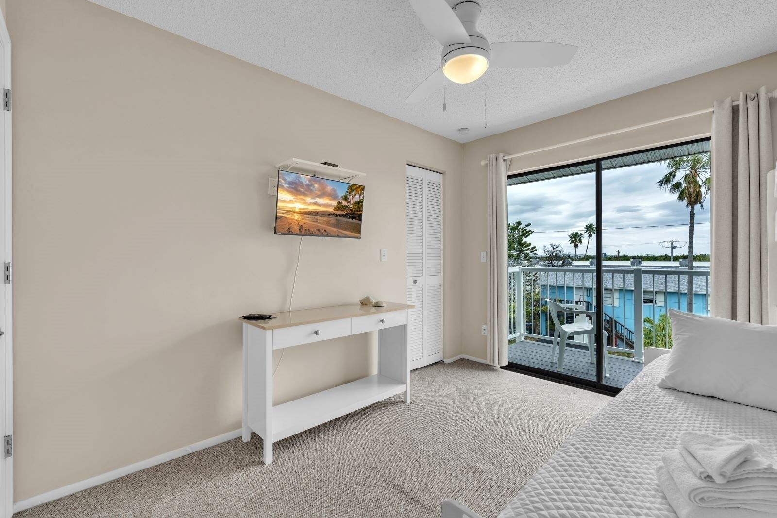 Quarto com cama, escrivaninha, cadeira, televisão e varanda com vista para uma piscina e palmeiras.