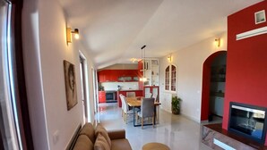 Un soggiorno moderno con un divano, un tavolo da pranzo e sedie. Si nota una cucina sullo sfondo, caratterizzata da mobili rossi e un camino.