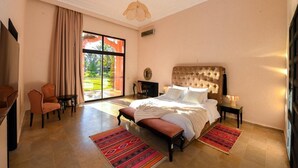 A spacious bedroom with a large bed, a sitting area with chairs, and a view through a sliding door to an outdoor area.