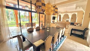 A spacious dining area with a long table and multiple chairs, large windows with a view of a garden, and traditional hanging lamps.