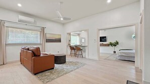A modern living room with a brown sofa, a dining area with two chairs, a small table, and a kitchen area with a TV and a plant.