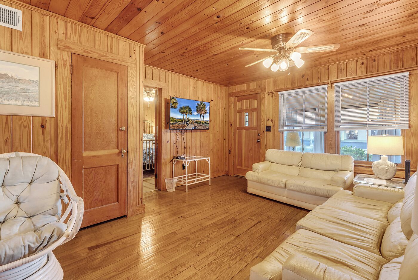Un intérieur en bois avec un ventilateur de plafond, un canapé blanc, un fauteuil en osier et un téléviseur fixé au mur.