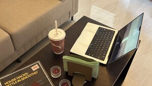 Een hotelkamer met een laptop op een houten tafel, een bank met een kussen, een tas, een smartphone en een boekje met de titel 'House Hacks: Comfortabel installeren'.