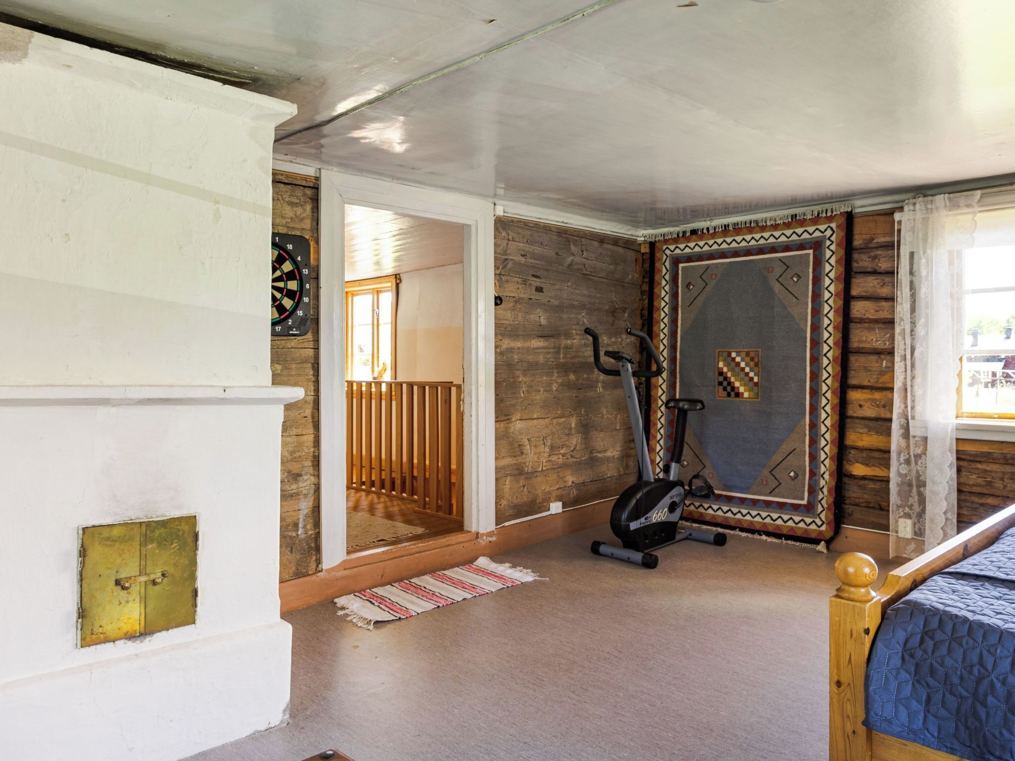 A room with a wooden floor, a bed with a blue quilt, a stationary exercise bike, a dartboard, and a door leading to a balcony.