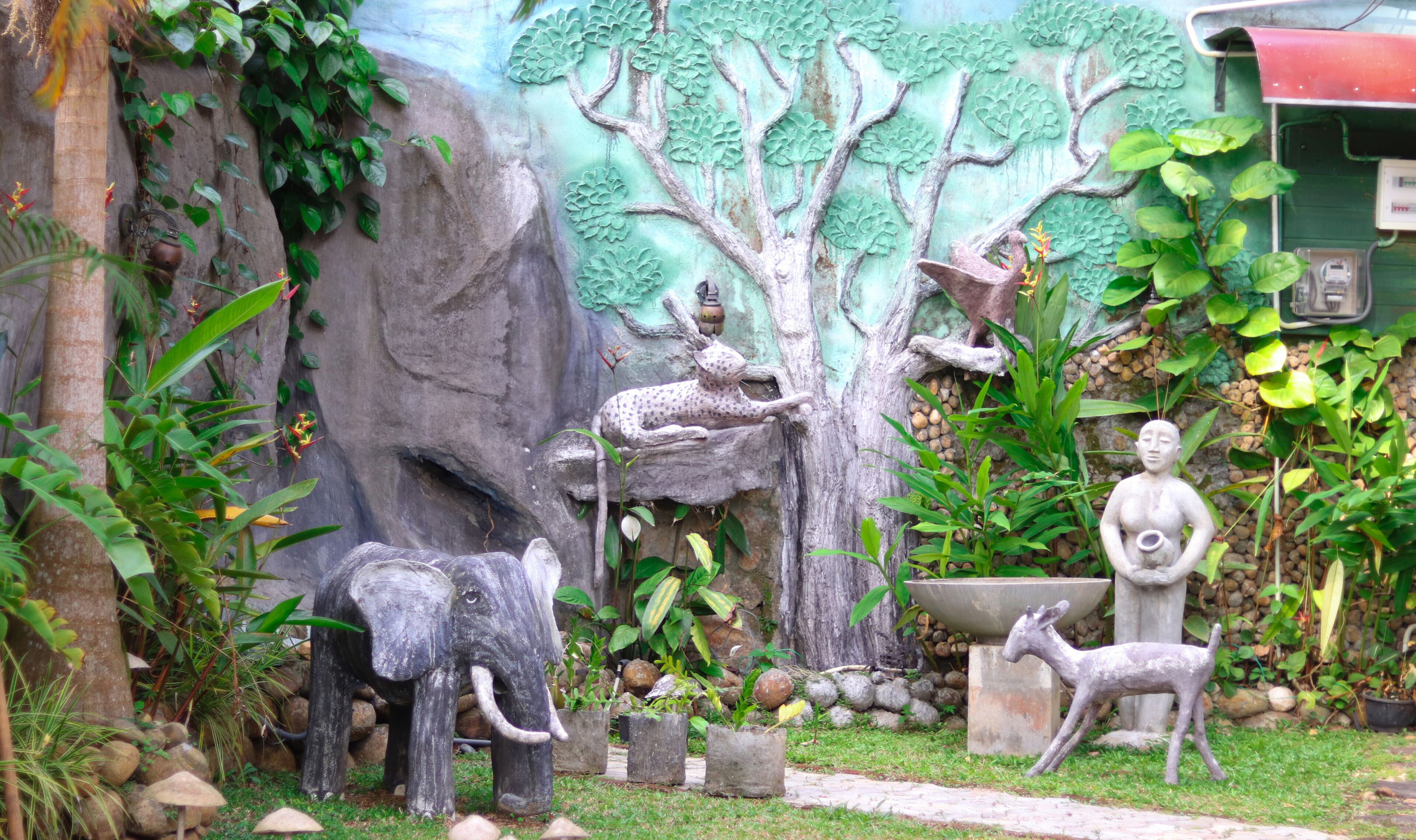 A garden with a rock wall, a tree sculpture, and a statue of a figure holding a cup.