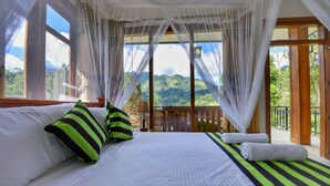 A bedroom with a large bed, a balcony with a wooden railing, and a view of lush greenery.