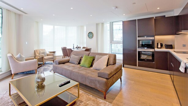 En moderne stue med en beige sofa, et glasbord og et køkken med mørke skabe og en mikrobølgeovn.