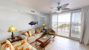 A living room with a sofa, two side tables, a coffee table, a ceiling fan, a fish wall decoration, and a large window with a view of the beach. - Beachfront Condo w/ Free round of Golf & Activities Daily! (Panama City Beach)
