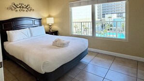 A hotel room with a large bed, a nightstand with a lamp, a decorative wall piece, and a window with a view of a pool and buildings. - Beachfront Condo w/ Free round of Golf & Activities Daily! (Panama City Beach)