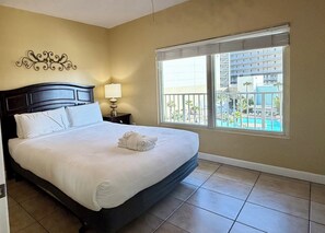 Una habitación de hotel con una cama grande, una mesita de noche con una lámpara, un adorno de pared y una ventana con vistas a una piscina y edificios.
