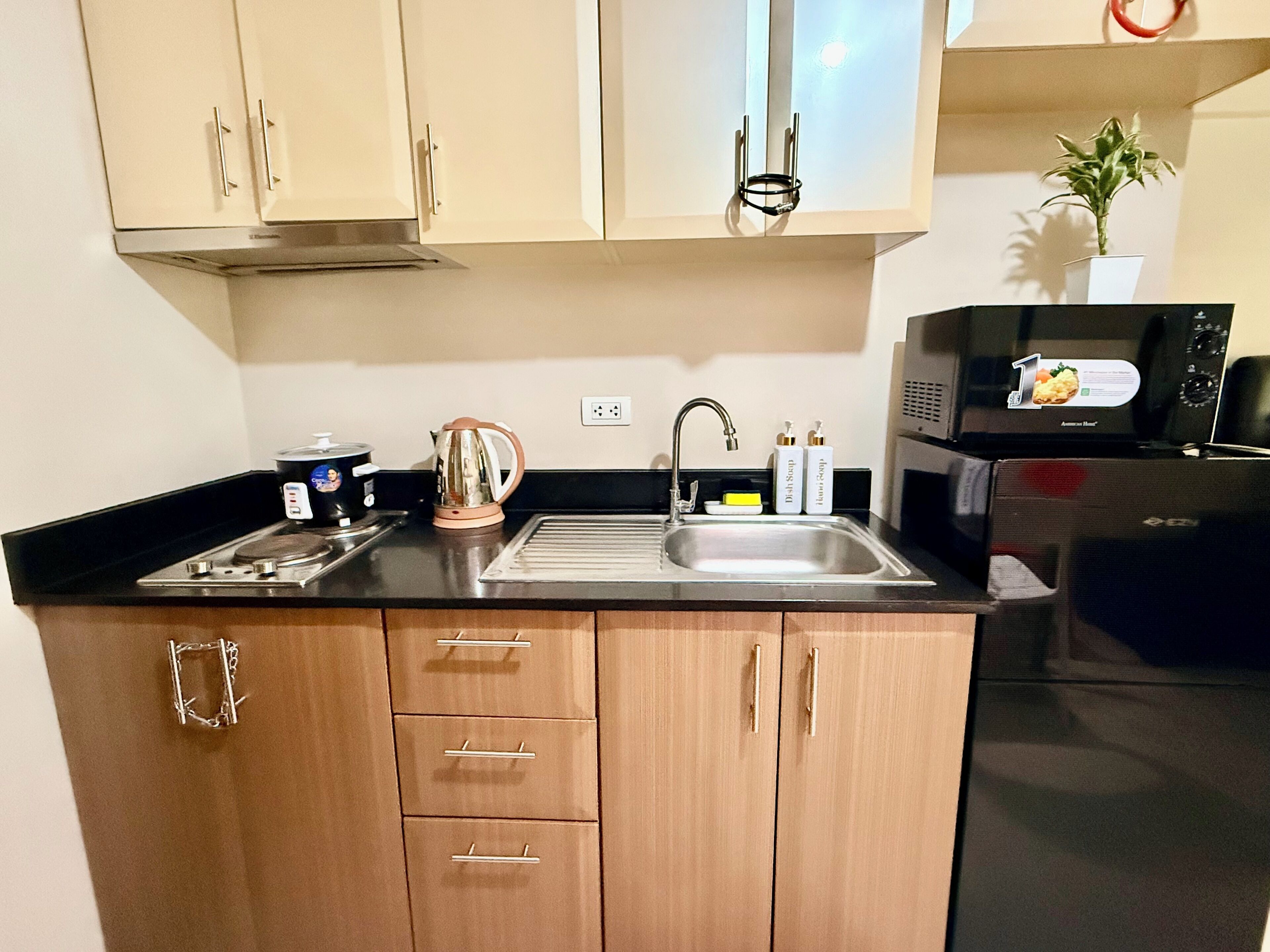 Una cocina compacta con armarios de madera, un lavamanos de acero inoxidable, un hervidor y un microondas.