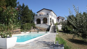 Área de piscina com piso de pedra, planta em vaso branco e um edifício com janelas em arco e varanda.