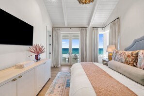 A bedroom with a bed, a TV, a nightstand, a lamp, a window with curtains, and a view of the ocean. - Beach Oasis | Luxury Gulf-Front Home with Private Heated Pool, Elevator & Beach (Pensacola Beach)