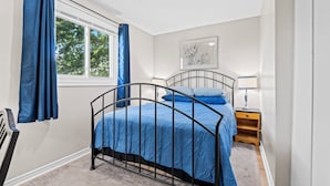 A bedroom with a metal bed frame, a window with blue curtains, a bedside table with a lamp, and a framed picture on the wall. - Cozy, Quiet Home in Kingston. Short drive to Queen's University. Pet Friendly! (Kingston)