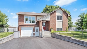 A two-story brick house with a white garage door, a red front door, and a small front yard with a white picket fence and a tree. - Cozy, Quiet Home in Kingston. Short drive to Queen's University. Pet Friendly! (Kingston)