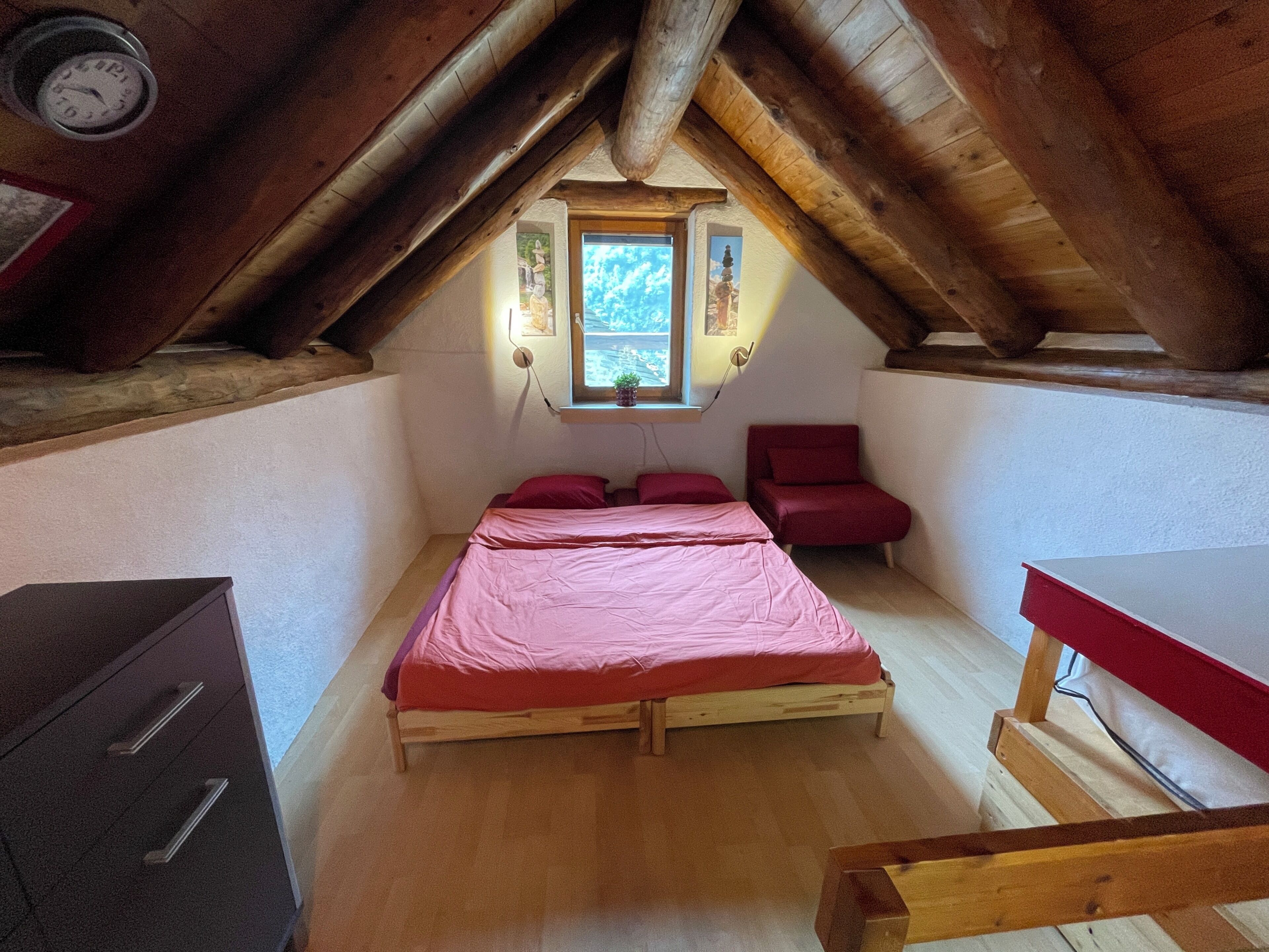 Een kleine, gezellige slaapkamer op zolder met een houten plafond, een enkel bed met rode beddengoed, een klein bureau en een raam dat natuurlijk licht binnenlaat.