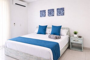 Ein ordentlich bezogenes Bett mit blauer und weißer Bettwäsche, ein kleiner Nachttisch mit einer Pflanze und drei Blumenbilder an der Wand.