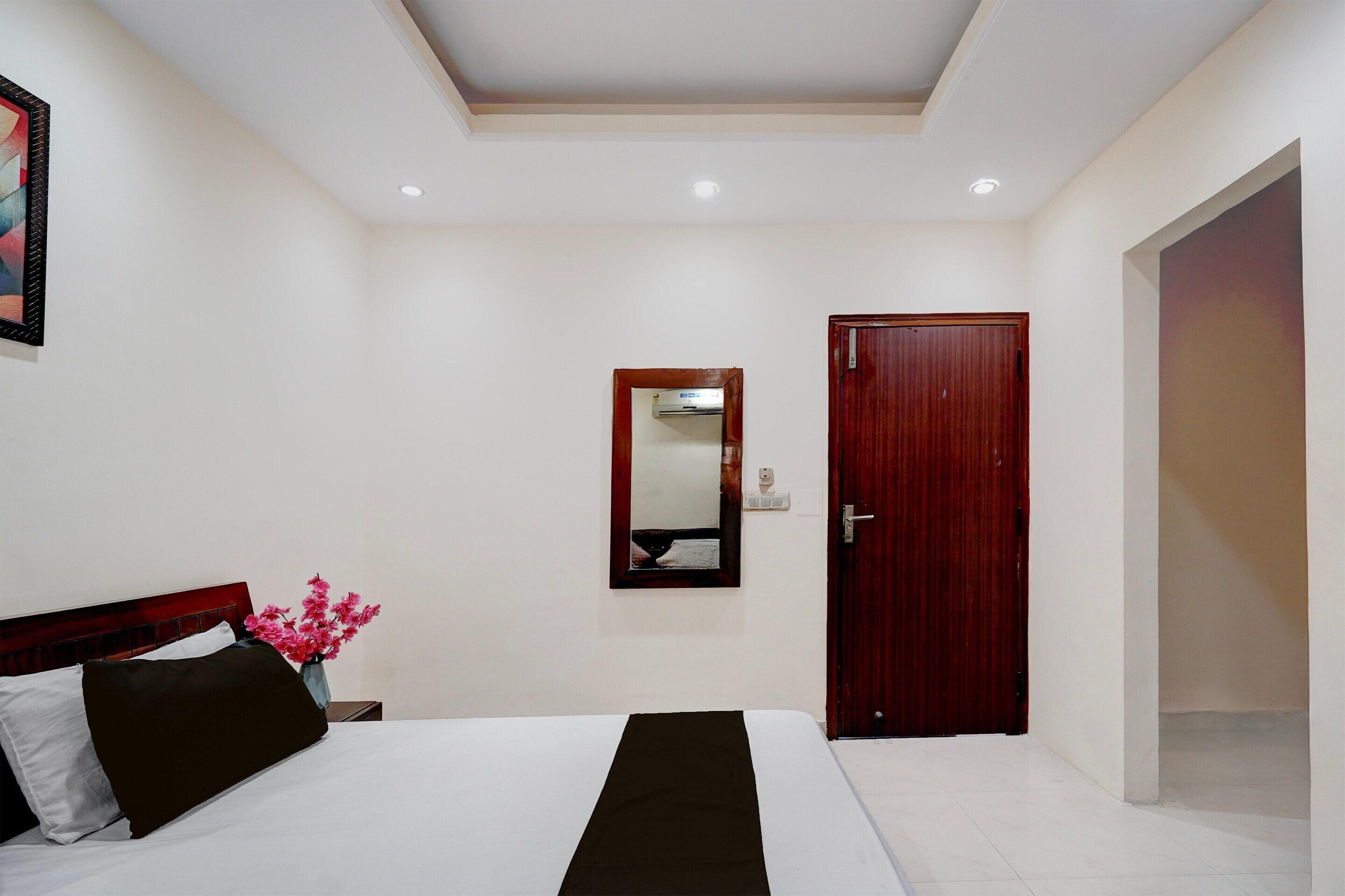 A hotel room with a bed, a wooden door, a mirror, and a nightstand with a vase of flowers.