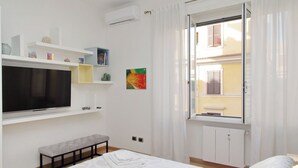 A bedroom with a bed, a bench, a TV, shelves with decorative items, and a window with curtains. - Casa Lenilli - Beautiful apartment in Testaccio (Roma)