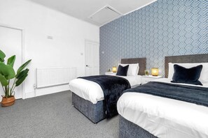 Ein Hotelzimmer mit zwei Betten, einer Wand mit Muster, einem Heizkörper, einer Zimmerpflanze und einem Nachttisch mit Lampe.