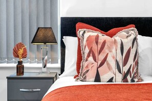 A hotel room with a bed, a nightstand with a lamp, a vase with dried flowers, and a window with curtains.