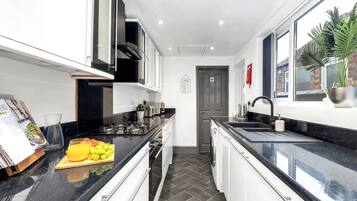 A modern kitchen with black countertops, white cabinets, and a black backsplash. There is a fruit bowl with grapes and an orange on the counter, and a potted plant on the windowsill.