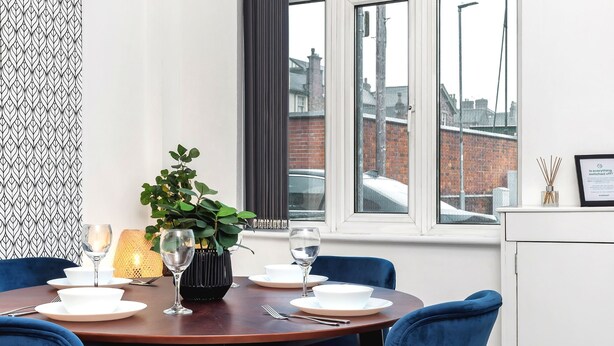 A dining area with a round table set for four, blue chairs, a potted plant, and a window with a view of buildings.