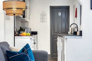 A compact living space with a kitchenette, a grey sofa with blue cushions, a hanging lamp, and a door with a sign indicating kitchen hours. - Chorlton House | Free Parking | 2BR Stoke-on-Trent (Stoke-on-Trent)