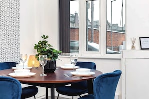 A dining area with a round table set for four, a potted plant, and a view of a brick building through the window. - Chorlton House | Free Parking | 2BR Stoke-on-Trent (Stoke-on-Trent)