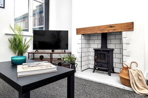 A modern living room with a flat-screen TV, a black wood-burning stove, a coffee table with books, and a potted plant.