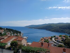 Eine Küstenstadt mit Häusern mit Terrakotta-Dächern, einem Hafen voller Boote und weiter Aussicht auf das Meer und sanfte Hügel in der Ferne.