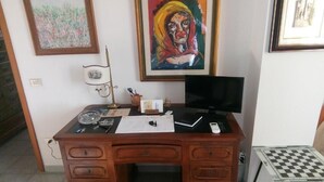 A wooden desk with a computer, papers, and a telephone. There are two paintings on the wall, a chair in front of the desk, and a small table with a checkered top to the right. - AT THE OLD STUDIO (Vibo Valentia)