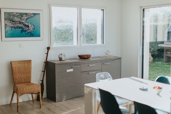 Una cocina moderna con comedor, que cuenta con un gran ventanal, una silla de mimbre, una mesa de madera y un armario con un bol encima.