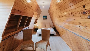 Un dormitorio acogedor en el ático con paredes de madera, una cama, dos sillones de mimbre y una mesita de noche con una lámpara.