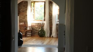 Ein Innenraum mit einer Steinsäule, einem Holzstuhl, einer großen grünen Vase und einem Fenster mit Vorhängen.