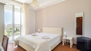 A hotel room with a large bed, a small bedside table with a lamp, a TV, and a balcony with a view of the city. - Panoramic Rooms  (Syracuse)