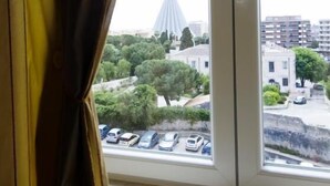 A view through a window with curtains, featuring a parking lot, a white building, and a distinctive tower in the background. - Panoramic Rooms  (Syracuse)