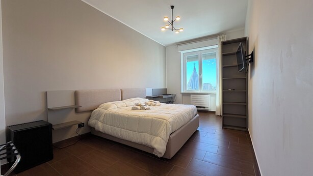 A bedroom with a bed, a desk, a chair, a radiator, a window with a city view, and a ceiling light fixture. - Panoramic Rooms  (Syracuse)