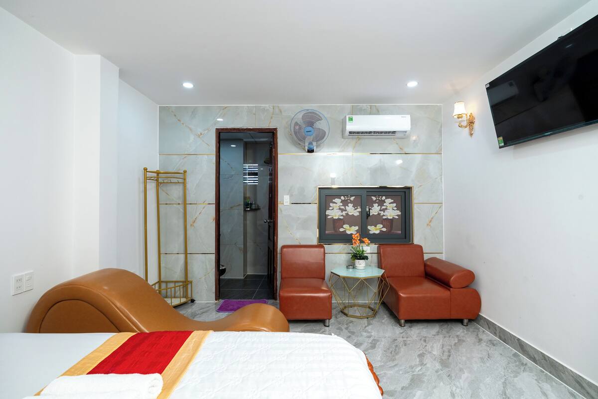 A hotel room with a bed, two leather armchairs, a small table with a vase, a wall-mounted TV, and an air conditioning unit.